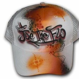 Peut inclure: Casquette de camionneur en maille blanche avec un design a&eacute;rographe orange, noir et blanc. Le design pr&eacute;sente les mots "Joe the Flo" en lettres noires.