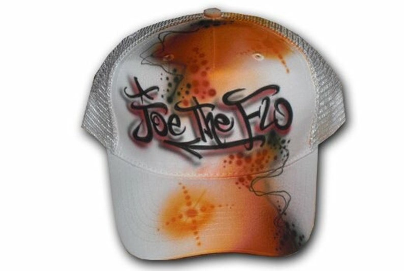 Peut inclure: Casquette de camionneur en maille blanche avec un design a&eacute;rographe orange, noir et blanc. Le design pr&eacute;sente les mots "Joe the Flo" en lettres noires.