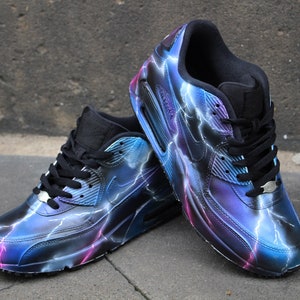 May include: A pair of black Nike Air Max 90 sneakers with a custom lightning bolt design. The sneakers are painted in shades of blue, purple, and pink.