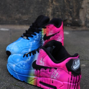 May include: A pair of custom-painted Nike Air Max 90 sneakers. One shoe is painted in a blue and yellow lightning bolt design, while the other is painted in a pink and black drip design. Both shoes feature a black swoosh logo.