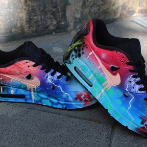 Puede incluir: Un par de zapatillas Nike Air Max 90 pintadas a mano con un esquema de colores vibrante. Las zapatillas tienen una base negra con salpicaduras de pintura rosa, azul, verde y blanca. El logotipo de Nike swoosh est&aacute; pintado en un color rosa claro.