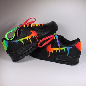 Puede incluir: Un par de zapatillas Nike Air Max 90 negras con pintura de goteo arco&iacute;ris y cordones de colores.