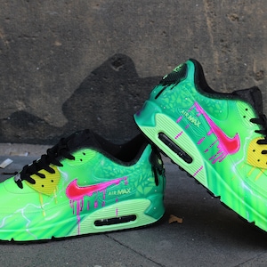 Puede incluir: Un par de zapatillas Nike Air Max 90 verdes con detalles en rosa y amarillo. Las zapatillas tienen un dise&ntilde;o de salpicaduras de pintura verde y negra con un patr&oacute;n de rayo.