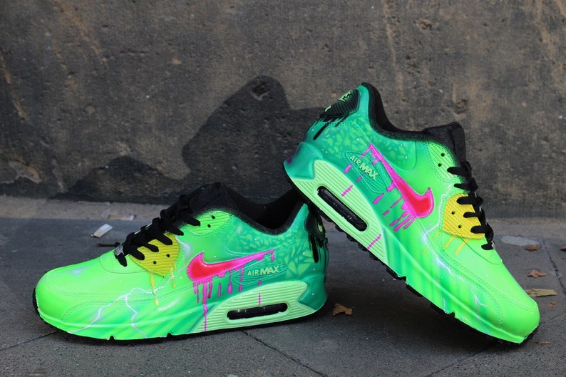 Puede incluir: Un par de zapatillas Nike Air Max 90 verdes con detalles en rosa y amarillo. Las zapatillas tienen un dise&ntilde;o de salpicaduras de pintura verde y negra con un patr&oacute;n de rayo.