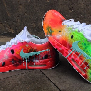 May include: A pair of custom-painted Nike Air Max 1 trainers with a vibrant red, orange, yellow, green, and blue colour scheme. The trainers feature a drip effect and lightning bolt design.