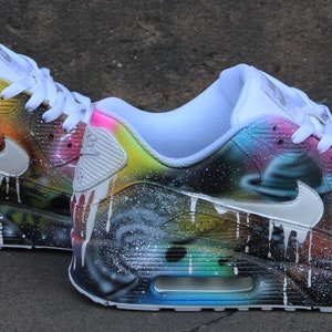 Op de afbeelding: Een paar witte Nike Air Max 90 sneakers met een galaxy-thema ontwerp. De sneakers hebben een zwart-witte achtergrond met kleurrijke accenten in blauw, groen, geel, oranje en roze. Het ontwerp bevat een galaxy print met sterren, planeten en nevels.