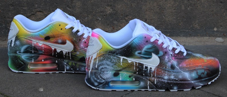 Op de afbeelding: Een paar witte Nike Air Max 90 sneakers met een galaxy-thema ontwerp. De sneakers hebben een zwart-witte achtergrond met kleurrijke accenten in blauw, groen, geel, oranje en roze. Het ontwerp bevat een galaxy print met sterren, planeten en nevels.