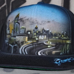 Könnte beinhalten: Eine schwarze und blaue Baseballkappe mit einem handgemalten Stadtbild. Das Stadtbild zeigt Gebäude, Straßen und Straßenlaternen. Die Kappe hat einen schwarzen Schirm und eine grüne Unterseite. Das Wort "Georgia" ist in Blau an der Seite der Kappe geschrieben.