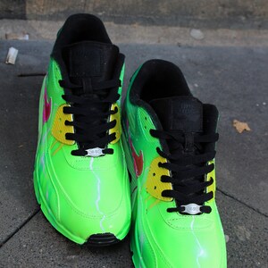 Puede incluir: Un par de zapatillas Nike Air Max 90 verdes con cordones negros y detalles amarillos. Las zapatillas tienen un dise&ntilde;o de rayo pintado en ellas.