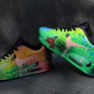 Puede incluir: Un par de zapatillas Nike Air Max 90 pintadas a mano. Una zapatilla es verde, amarilla y naranja con un swoosh rosa. La otra zapatilla es verde, azul y rosa con un swoosh blanco. Ambas zapatillas tienen cordones negros y detalles negros.