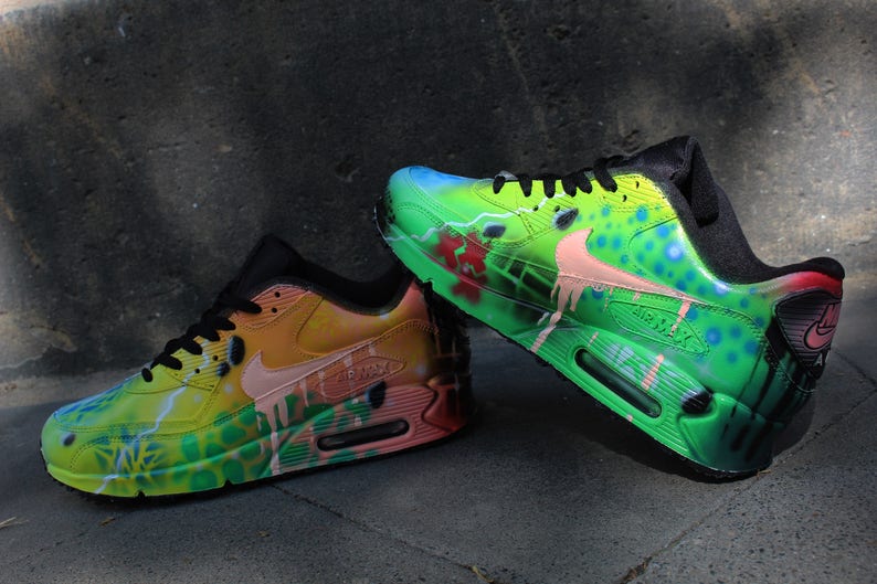 Puede incluir: Un par de zapatillas Nike Air Max 90 pintadas a mano. Una zapatilla es verde, amarilla y naranja con un swoosh rosa. La otra zapatilla es verde, azul y rosa con un swoosh blanco. Ambas zapatillas tienen cordones negros y detalles negros.