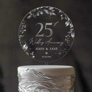 May include: A silver cake topper with a floral design and the text "25th Wedding Anniversary John & Jane 09.18.1999".