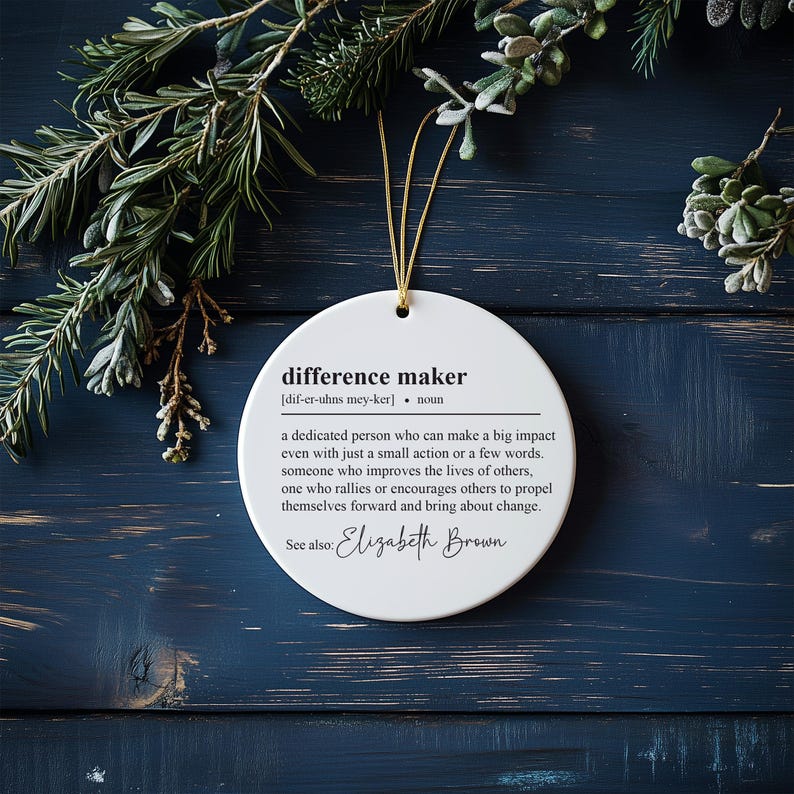 May include: White round bauble with the definition of "difference maker" and the name "Elizabeth Brown" printed in black text.