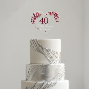 May include: A white cake with silver leaf accents topped with a clear heart-shaped acrylic cake topper. The topper features a burgundy floral design and the text "40th Wedding Anniversary John & Jane 05.10.1984".