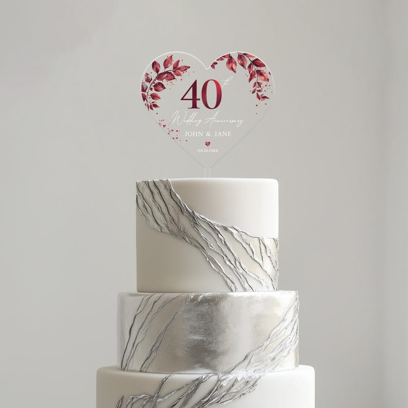 May include: A white cake with silver leaf accents topped with a clear heart-shaped acrylic cake topper. The topper features a burgundy floral design and the text "40th Wedding Anniversary John & Jane 05.10.1984".
