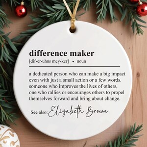 May include: White round bauble with the definition of "difference maker" and the name "Elizabeth Brown" printed on it. The bauble is hanging on a Christmas tree.