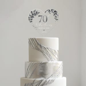 Op de afbeelding: Een witte bruidstaart met zilveren accenten en een hartvormige, transparante taarttopper met de tekst "70e huwelijksverjaardag John & Jane 09.18.1954".