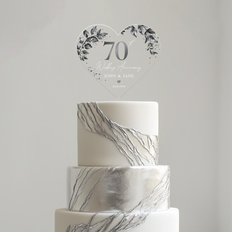 Op de afbeelding: Een witte bruidstaart met zilveren accenten en een hartvormige, transparante taarttopper met de tekst "70e huwelijksverjaardag John & Jane 09.18.1954".