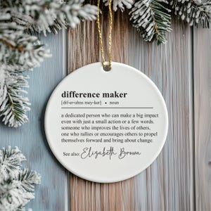 May include: White ceramic bauble with the definition of "difference maker" and the name "Elizabeth Brown" printed in black.