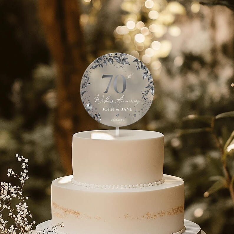 Op de afbeelding: Een witte tweelaagse taart met een ronde, zilveren taarttopper waarop staat "70e huwelijksverjaardag John & Jane 09.18.1954". De taarttopper heeft een aquarelontwerp met blauwe bladeren en bloemen.