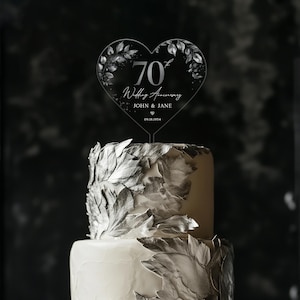 Op de afbeelding: Een transparante, hartvormige taarttopper van acryl met een zilveren glitterontwerp en de tekst "70e huwelijksverjaardag John & Jane 09.18.1954".