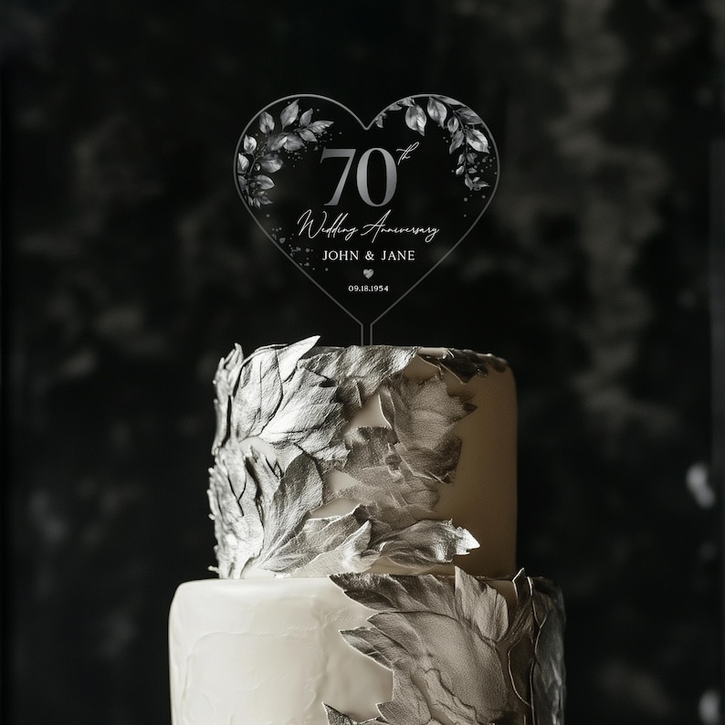 Op de afbeelding: Een transparante, hartvormige taarttopper van acryl met een zilveren glitterontwerp en de tekst "70e huwelijksverjaardag John & Jane 09.18.1954".