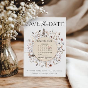 Puede incluir: Tarjeta de guardar la fecha con un dise&ntilde;o de corona floral. La tarjeta presenta un c&iacute;rculo con un calendario para abril de 2024 con la fecha 12 marcada en rojo. El texto "Save the Date" est&aacute; en la parte superior de la tarjeta y "Amanda & David" est&aacute; debajo del calendario. El texto "Marigold Hotel, Santa Rosa, California" y "Invitaci&oacute;n formal a seguir" est&aacute;n en la parte inferior de la tarjeta.