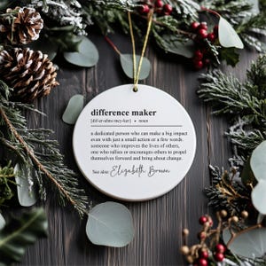 May include: White round Christmas bauble with black text defining "difference maker" as a dedicated person who can make a big impact. The bauble also includes the text "See also: Elizabeth Brown".