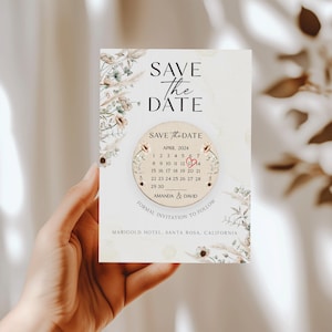 Puede incluir: Tarjeta de guardar la fecha con un dise&ntilde;o floral y un c&iacute;rculo de madera con un calendario que muestra abril de 2024 con un coraz&oacute;n marcando el 13. El texto dice "Save the Date" y "Amanda & David" con la ubicaci&oacute;n "Marigold Hotel, Santa Rosa, California".