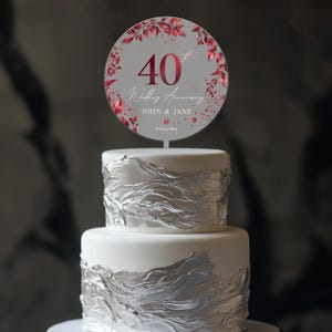 May include: A round cake topper with a grey background and red floral accents. The topper features the number 40, the text "Wedding Anniversary", the names "JOHN & JANE", and the date "09.18.1984".
