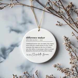 May include: White round ornament with the definition of "difference maker" and the name "Elizabeth Brown" printed in black text.