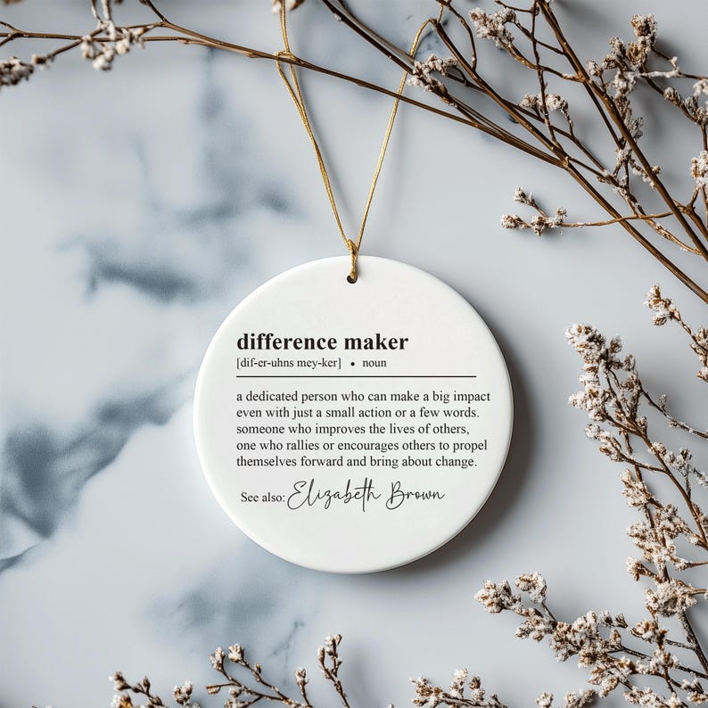 May include: White round ornament with the definition of "difference maker" and the name "Elizabeth Brown" printed in black text.