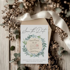 Puede incluir: Tarjeta de guardar la fecha con una corona de acuarela de hojas verdes y un c&iacute;rculo de calendario con un coraz&oacute;n rojo marcando el 12 de abril de 2024. El texto dice "Save the Date" y "Amanda & David".