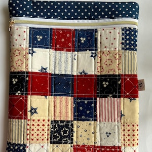 May include: A red, white and blue quilted zippered pouch with a patchwork design featuring stars and stripes. The pouch is made of fabric and has a gold zipper.