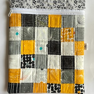 Peut inclure: Une pochette matelassée avec un motif patchwork. Les carrés présentent des motifs jaunes, gris, noirs et blancs. Une fermeture éclair blanche et un dessus à motifs floraux complètent le design. La pochette mesure environ 25 cm de large et 30 cm de haut.