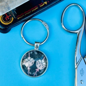 May include: A silver keyring with a circular pendant displaying a floral pattern on a dark backdrop. The image is set against a light blue surface, with a sewing machine and scissors in the background.