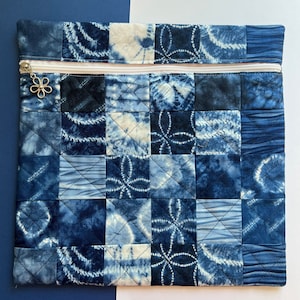 May include: A blue quilted zippered pouch with a patchwork design. The pouch is made of various shades of blue fabric with a white zipper and a silver flower charm.