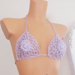 May include: A lavender crochet bikini set on a mannequin. The halter-neck top has floral detailing on the cups. The matching bottoms feature side ties and a floral design. The text "sinurgdesigns" is visible.
