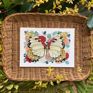 May include: A watercolour illustration of a butterfly with brown and white wings surrounded by pink, red and white flowers. The illustration is printed on white paper and framed in a wicker tray.