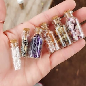 May include: Six small glass bottles with cork tops, each filled with different colored crystals and stones. The bottles are held in a person's hand.
