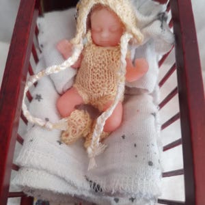 May include: A miniature baby doll in a wooden crib. The doll is wearing a knitted yellow romper, matching bonnet, and booties. The baby is lying on a white blanket with a floral patterned sheet underneath. The crib is dark red.