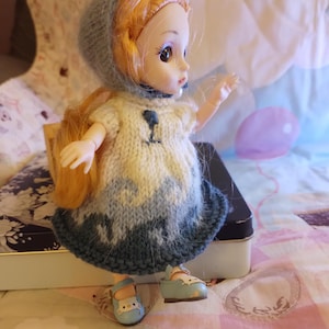 May include: A doll with red hair wearing a blue knitted hat, a white and blue knitted dress, and blue shoes. The doll is standing on a patterned quilt. The dress has a small blue design on the front.