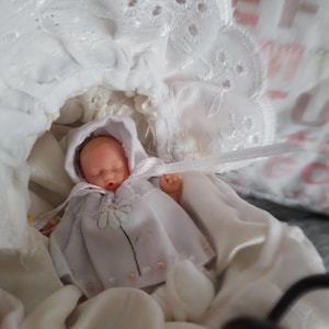 Op de afbeelding: Een miniatuurpop met een lichte huidskleur, gekleed in een wit outfit met capuchon, voorzien van een madeliefje en een wit lintje. De pop ligt in een witte stoffen wieg die is afgewerkt met een gedetailleerde broderierand.
