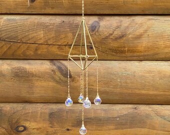 May include: A gold geometric mobile with five iridescent crystal drops hanging from it. The mobile is suspended from a chain and is hanging against a wooden background.