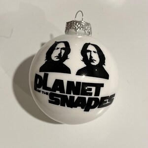 May include: White Christmas ornament with a black and white image of two faces and the text "Planet of the Snapes".