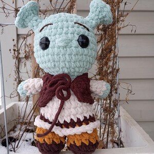 May include: A handmade crochet doll with a light blue head, large black eyes, and brown eyebrows. The doll wears a burgundy vest, a cream-colored dress with brown and yellow accents, and brown shorts. The doll has light blue arms and ears.
