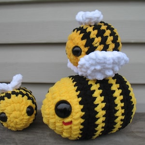 May include: Three crocheted bumblebees, one small, one medium, and one large, are stacked on top of each other. The bees are yellow and black with white wings and black eyes.