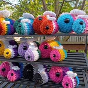 May include: A collection of colorful, handmade crochet bee plushies displayed on a black metal shelf. The bees feature a variety of colors, including rainbow, orange, blue, pink, and purple. Each bee has a large black eye and a white fluffy top.