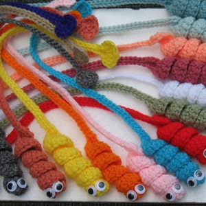 May include: A collection of colorful, crocheted worm-like toys with googly eyes. Each worm features a different color, including red, orange, yellow, green, blue, and pink. The toys are arranged on a white surface, showcasing their whimsical design.