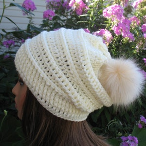 May include: Cream-colored, crocheted beanie hat with a large, fluffy, light-brown pom-pom. The hat has a ribbed texture and a slouchy fit. The background features green foliage and pink flowers.